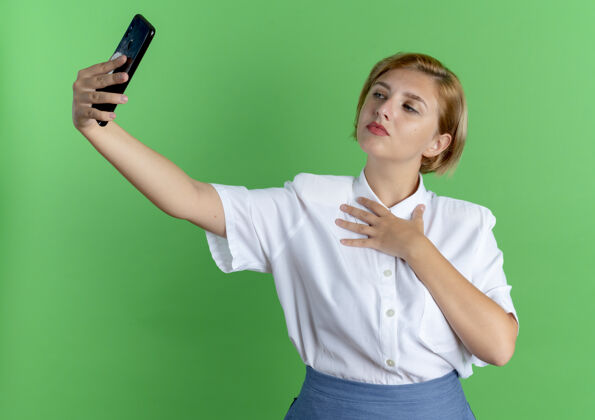 俄罗斯年轻自信的金发俄罗斯女孩看着手机自拍把手放在胸前隔离在绿色背景与复制空间胸部女孩自拍