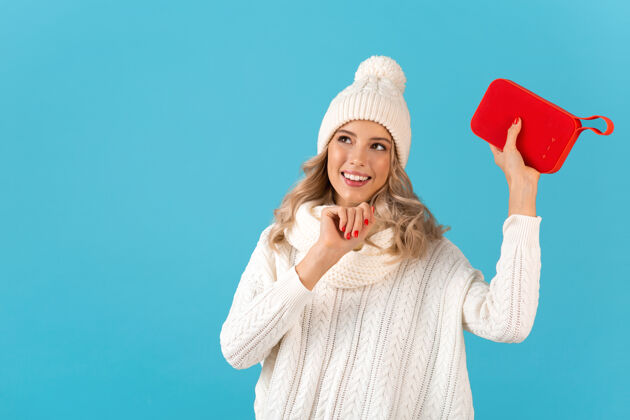 现代时尚的金发微笑美丽的年轻女子手持无线扬声器听音乐快乐的跳舞穿着白色毛衣和针织帽子冬季时尚的姿势隔离在蓝色的墙上穿着音频无线