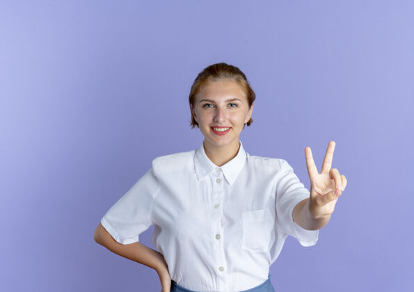 手年轻微笑的金发俄罗斯女孩手势胜利的手标志把手放在腰部孤立的紫色背景与复制空间手势女孩微笑
