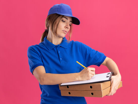 脸自信的年轻漂亮的女送货员穿着制服 拿着比萨饼盒 在剪贴板上写字 笔被隔离在粉红色的墙上制服公民递送