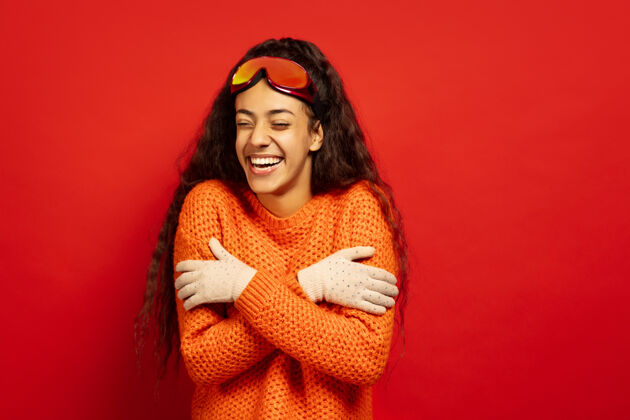 女性美国黑人年轻黑发女子的肖像滑雪面具上的红色工作室背景概念的人类情感 面部表情 销售 广告 冬季运动和假日在寒冷中温暖 笑声眼镜女孩成人