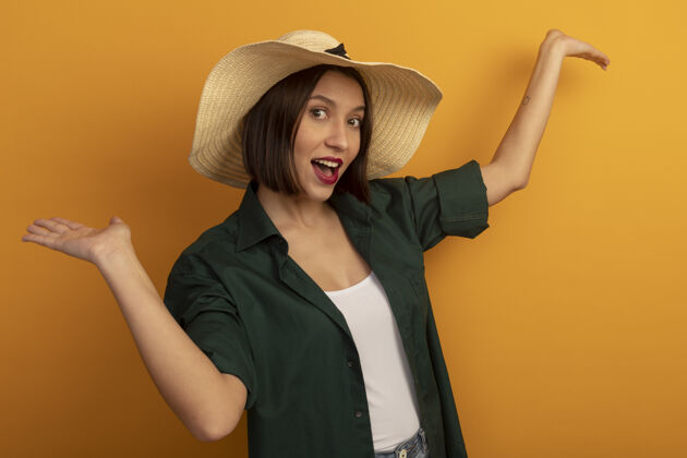 人快乐的漂亮女人戴着沙滩帽 举手站在橙色的墙上姿势快乐帽子