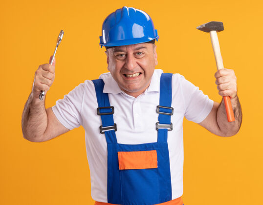 兴奋兴奋的成年建筑工人在制服举行扳手和锤子隔离在橙色墙上锤子举行表情