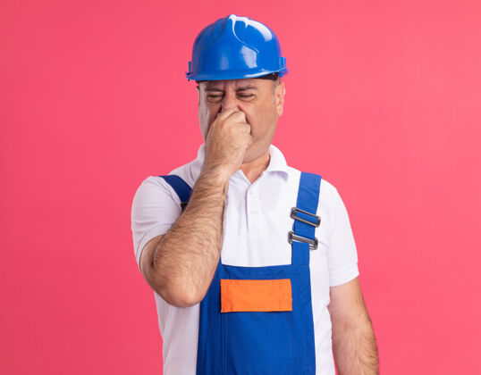 脸穿制服的未出租的成年建筑工人把鼻子隔离在粉红色的墙上不愉快人人