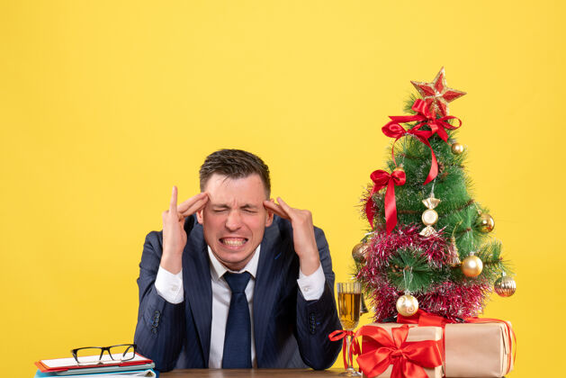 礼物前视图愤怒的男人把手指放在他的太阳穴上 坐在圣诞树旁的桌子上 黄色背景上的礼物坐西装太阳穴