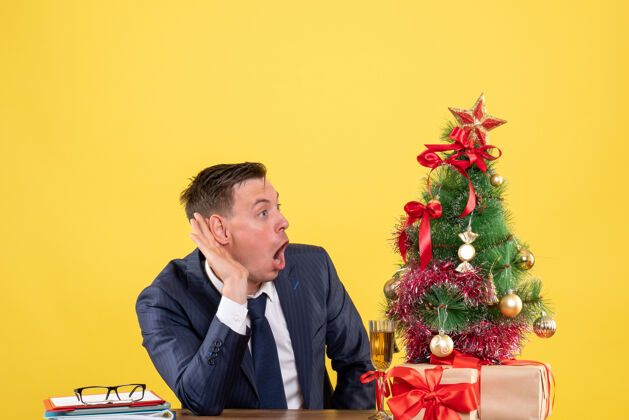 生意前视图令人惊讶的人坐在圣诞树旁的桌子旁 听着黄色背景上的礼物树新郎行政