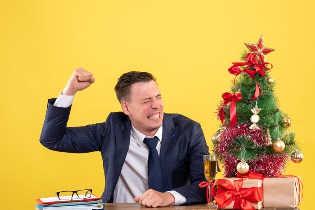 愤怒的男人前视图愤怒的男人举起他的手坐在圣诞树和黄色背景上的礼物旁边的桌子上会议男性生意