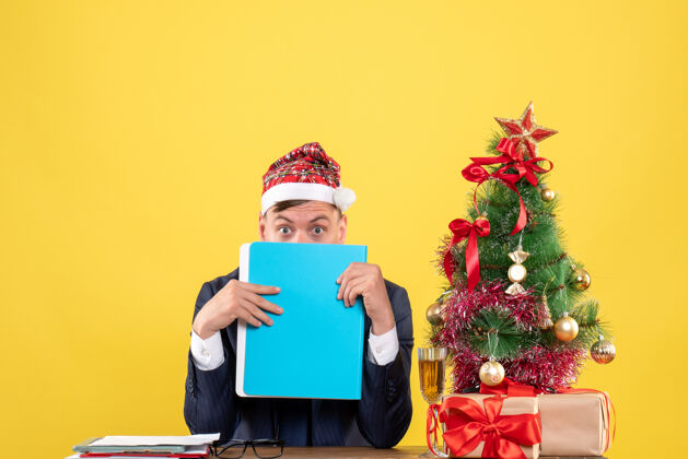 附近前视图商人用文件夹盖住他的脸 放在圣诞树旁的桌子上 黄色背景上的礼物前面桌子背景