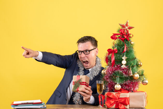 愤怒的人前视图愤怒的人戴着眼镜坐在圣诞树旁的桌子旁 黄色背景的礼物圣诞节桌子快乐