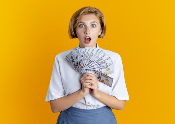 相机年轻的惊讶的金发俄罗斯女孩拿着钱看着相机孤立的橙色背景与复制空间钱金发女郎俄国人