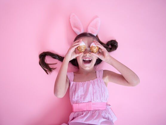 顶视图带着复活节兔子耳朵的小女孩在粉红色的墙上摆着复活节彩蛋复活节复活节彩蛋节日