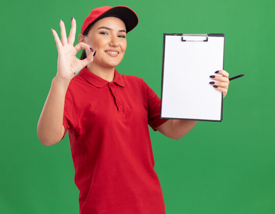 站着身穿红色制服 戴着帽子的年轻送货员拿着空白页的剪贴板 面带微笑地看着前面 绿色墙上立着“ok”的牌子展示拿着页面