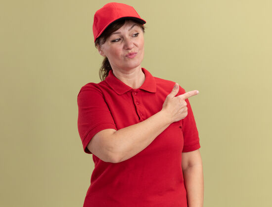 站立身穿红色制服 头戴鸭舌帽的中年女送货员自信地用食指指着站在绿色墙上的一边自信年龄制服