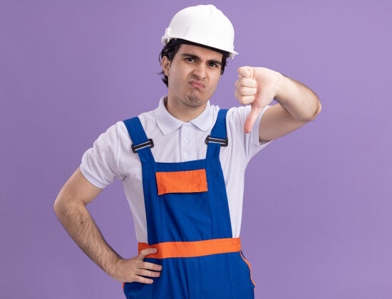 不高兴年轻的建筑工人穿着建筑制服 戴着安全帽 不高兴地看着前面 竖起大拇指站在紫色的墙上男人建筑展示