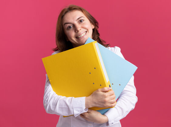 抱着身穿白衬衫的年轻女子拿着文件夹 站在粉红色的墙上 面带微笑地看着前面脸年轻衬衫