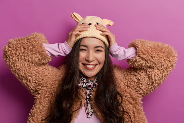 韩国人漂亮的亚洲女人的肖像很乐意买新帽子 手放在头上 真诚地微笑 穿着冬衣 露出洁白的牙齿情绪亚洲人墙