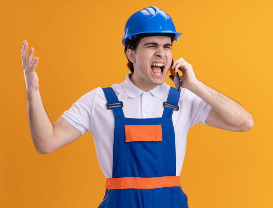 建设者身穿施工制服 头戴安全帽的年轻建筑工人站在橘红色的墙边 用咄咄逼人的表情大声喊叫着愤怒和疯狂电话喊叫制服