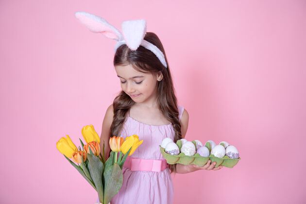 花有复活节兔子耳朵的小女孩手里拿着一束郁金香和一盘鸡蛋放在粉红色的墙上节日小托盘