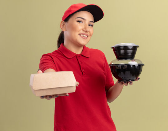 帽子身着红色制服 头戴鸭舌帽 面带微笑地站在绿色墙壁上 拿着食品包的快乐的年轻送货员望着前方年轻拿着食物