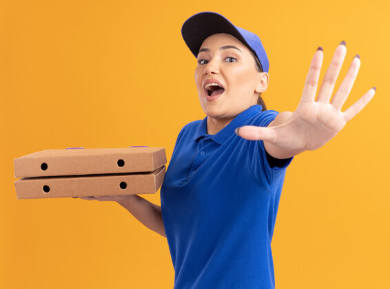 停止身穿蓝色制服 头戴鸭舌帽的年轻女送货员拿着比萨饼盒望着前面 站在橙色的墙上做着停下的手势 担心的手拿着站着女人