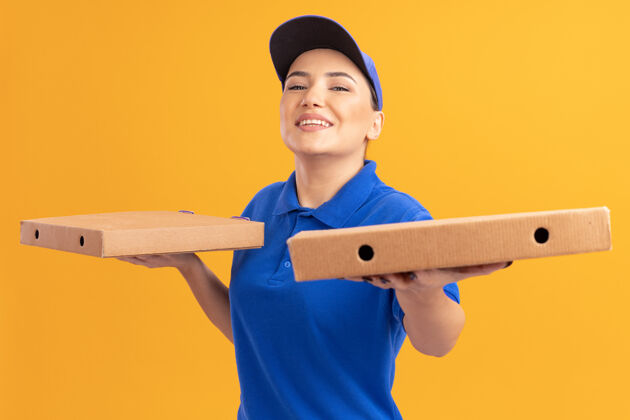 微笑身着蓝色制服 头戴鸭舌帽 手持披萨盒 站在橙色墙壁上 面带微笑 快乐地看着前方的年轻送货员制服送货披萨