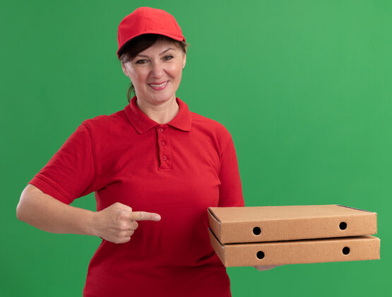 女人身穿红色制服 头戴鸭舌帽的中年女送货员拿着披萨盒 用食指指着盒子 微笑着自信地站在绿色的墙上帽子微笑制服
