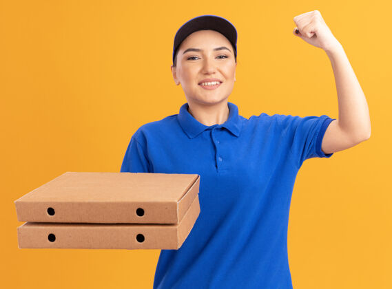 举行身穿蓝色制服 头戴鸭舌帽 拿着比萨饼盒的年轻女送货员微笑着自信地举起拳头看着前方 就像一个站在橙色墙壁上的胜利者信心盒子拳头