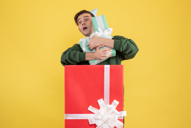 男正面图戴着派对帽的年轻人拿着礼物站在黄色背景的大礼盒后面帽子快乐礼盒