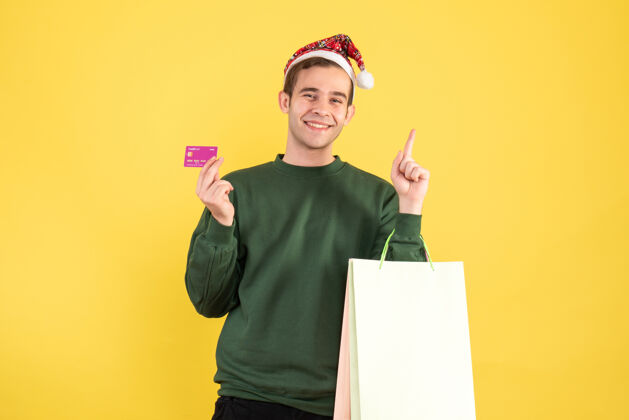前面正面图：戴着圣诞帽的年轻人拿着购物袋和购物卡站在黄色的背景复制空间上男人背景人