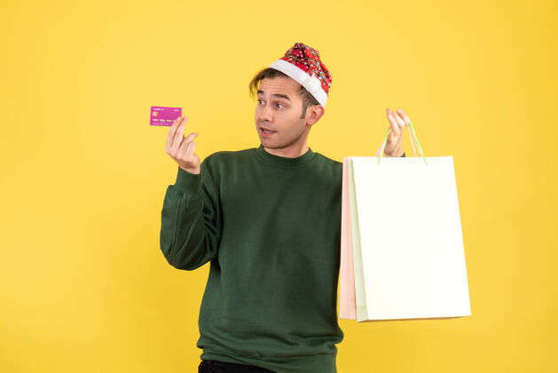 微笑正面图：一个戴着圣诞帽的年轻人拿着购物袋和购物卡站在黄色背景的地方包工作举行