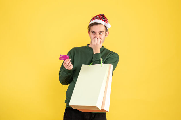 西装正面图：戴着圣诞帽的年轻人拿着购物袋和信用卡站在黄色背景上执行背景举行