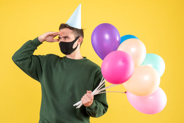 多彩正面图：站在黄色背景上观察戴着派对帽和彩色气球的年轻人欢乐庆祝聚会