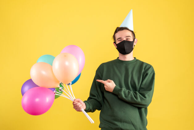 气球正面图：戴着派对帽的年轻人指着站在黄色背景上的彩色气球节日立场年轻人