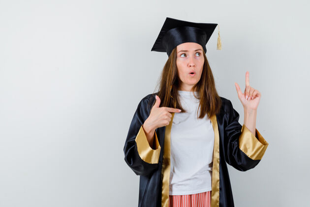 教育女毕业生的画像 穿着学院服 向上和右指 看着犹豫不决的前视图微笑帽子妇女