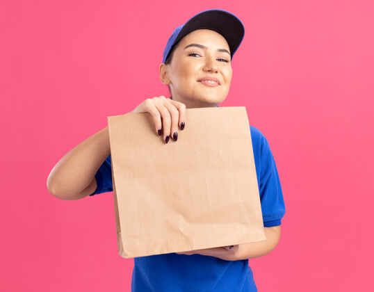 快乐快乐的年轻送货员 身穿蓝色制服 戴着帽子 手里拿着纸包 站在粉红色的墙上 面带微笑地看着前面站着制服递送
