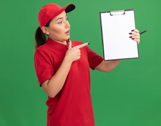 困惑身穿红色制服 戴着帽子的年轻女送货员拿着空白页的剪贴板 用食指指着剪贴板 站在绿色的墙上 神情困惑立场指向女人