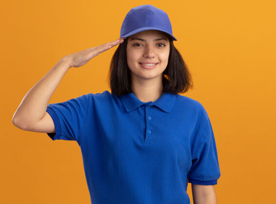 自信身着蓝色制服 头戴鸭舌帽的年轻送货员站在橙色的墙上微笑着自信地敬礼敬礼制服站立