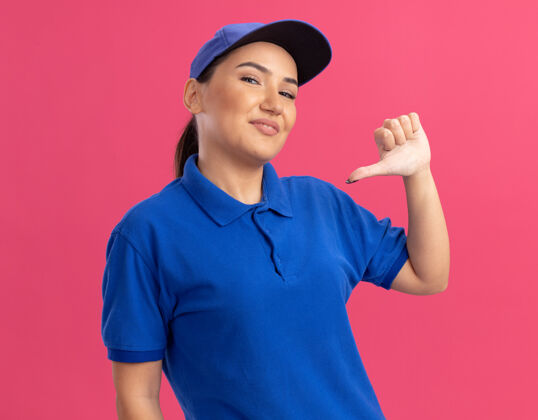 自信身穿蓝色制服 头戴鸭舌帽的年轻女送货员高兴而自信地用拇指指着站在粉红色墙上的自己帽子分娩指点