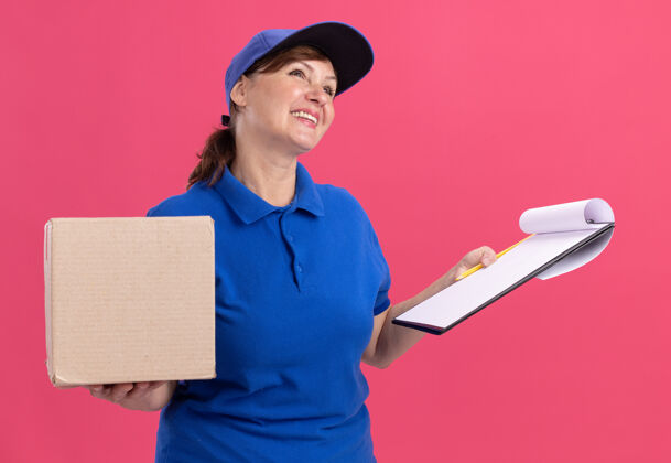 拿着身穿蓝色制服 头戴鸭舌帽的中年女送货员站在粉红色的墙上 手里拿着纸板箱和空白页的剪贴板 脸上带着微笑抬起头来空白递送制服