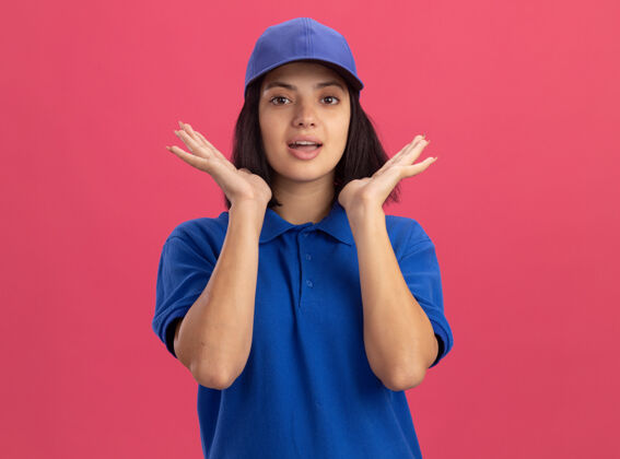 举起身穿蓝色制服 头戴鸭舌帽的年轻送货员站在粉红色的墙上 惊讶地举起双臂惊人女孩帽子