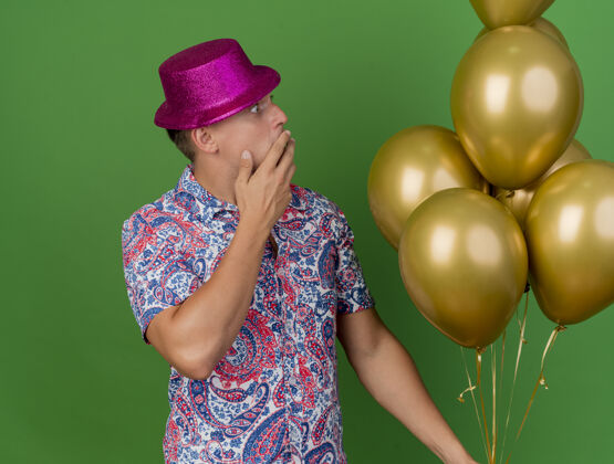 掩护被吓坏了的年轻人戴着粉红色的帽子拿着气球看着满嘴的手孤立在绿色的背景上手戴着嘴