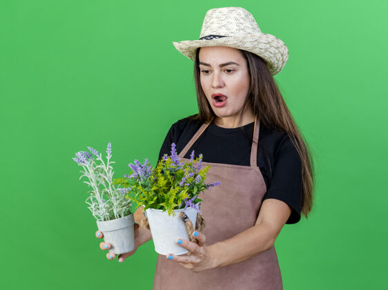 园艺一个穿着制服的美丽的园丁女孩戴着园艺帽 手拿花盆 看着隔离在绿色背景上的花朵花穿举行