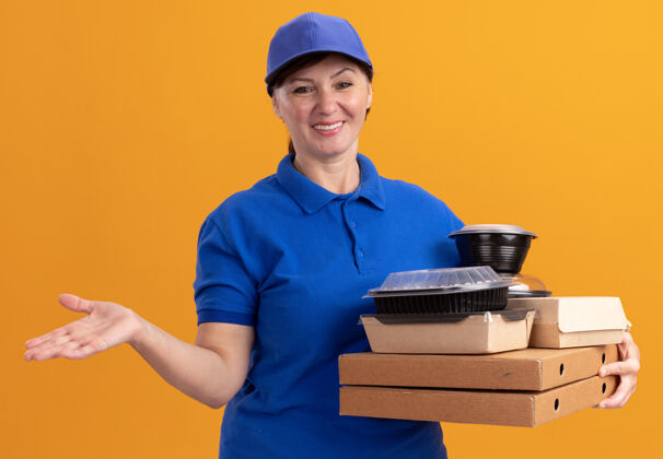 帽子快乐的中年送货员身穿蓝色制服 戴着帽子 手里拿着披萨盒和食品包 站在橘色的墙上 面带微笑地看着前面女人披萨制服