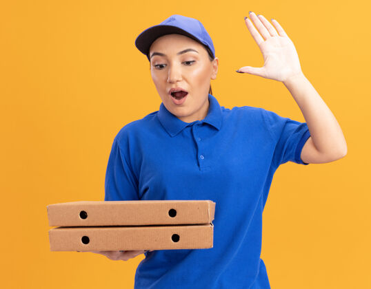 惊喜身穿蓝色制服 头戴鸭舌帽 手持比萨饼盒的年轻送货妇女站在橙色的墙上 惊讶地举手看着比萨饼盒站着盒子提高