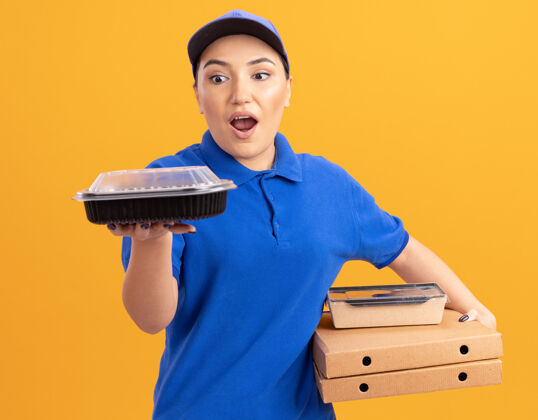 制服身穿蓝色制服 头戴鸭舌帽的年轻女送货员拿着比萨饼盒和食品包 站在橘色的墙上惊讶地看着盒子女人帽子站着