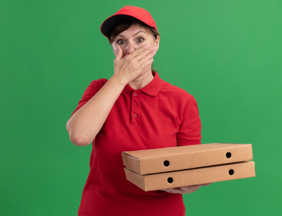 年龄身穿红色制服 头戴鸭舌帽的中年女送货员拿着披萨盒望着前面 震惊地捂着嘴站在绿色的墙上手封面女人