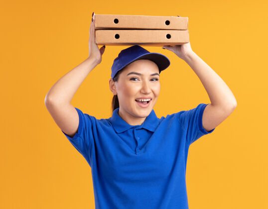 抱着身着蓝色制服 头戴鸭舌帽的快乐的年轻送货女 头上举着披萨盒 微笑自信地站在橙色的墙上看着前方站着开心女人