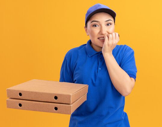 制服身穿蓝色制服 头戴鸭舌帽的年轻女送货员拿着比萨饼盒 看着站在橘色墙壁上紧张不安 咬牙切齿的钉子紧张盒子披萨