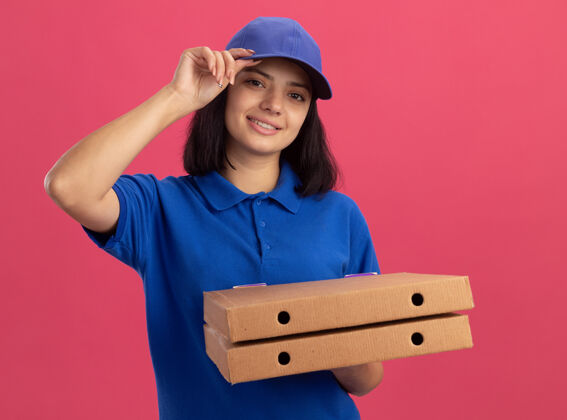 触摸身穿蓝色制服 戴着披萨盒的年轻送货员微笑着 自信地抚摸着站在粉色墙上的帽子微笑帽子制服