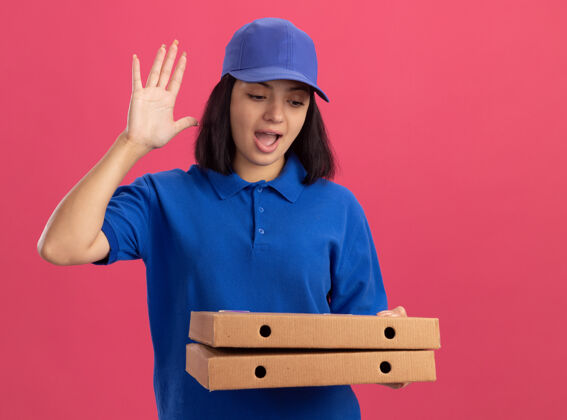 制服身穿蓝色制服 头戴鸭舌帽 拿着比萨饼盒的年轻送货员快乐而积极地举起手掌站在粉红色的墙上举起拿着年轻